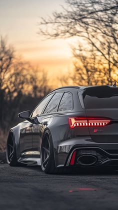 the rear end of a grey car parked in front of some trees at sunset with its lights on