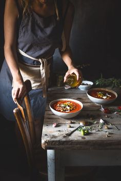 a woman pours olive oil into two bowls of soup on a wooden table in front of her