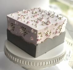 a square cake with pink and grey frosting on a white plate next to a window