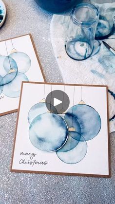 two cards with watercolor ornaments hanging from them on a table next to some glassware