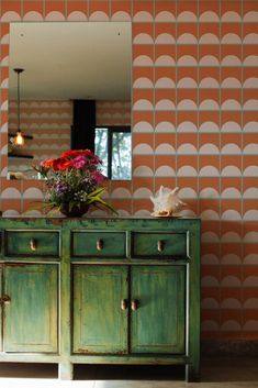 a green dresser with flowers on top in front of a wallpapered mirror and lamp