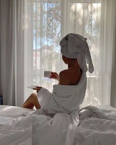 a woman sitting on a bed with a towel wrapped around her head and holding a cup