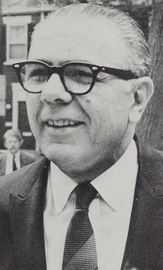 black and white photograph of a man in suit and tie with glasses smiling at the camera