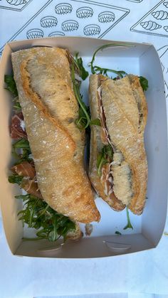 two sandwiches in a white box on top of a paper wrapper with green leaves