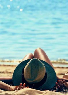 a woman laying on top of a sandy beach next to the ocean wearing a hat