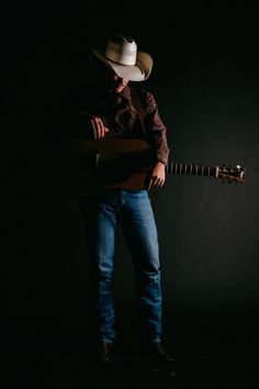 a man wearing a cowboy hat and holding a guitar in his right hand while standing against a black background