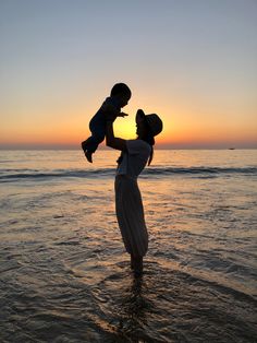 a woman holding a child in her arms while standing in the ocean at sunset or sunrise