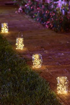 lighted mason jars lined up on the side of a brick path at night with flowers in the background