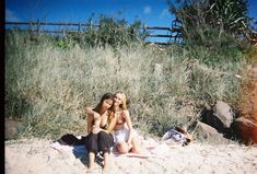 two young women sitting on the sand in front of some tall grass and bushes, posing for a photo