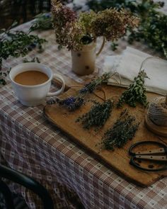 a wooden cutting board topped with lots of herbs next to a cup of coffee and scissors