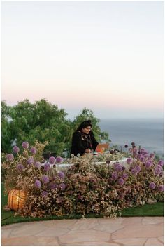 a woman sitting at a table surrounded by purple flowers and greenery with the ocean in the background