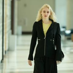 a woman with blonde hair walking down a hall wearing a black suit and yellow shirt