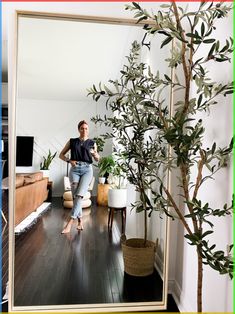 a woman standing in front of a mirror next to a potted tree and couch