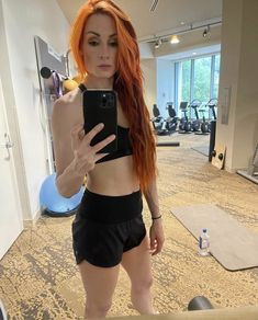 a woman with red hair taking a selfie in a gym room while holding a cell phone