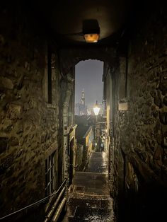 an alley way with stone walls and buildings in the background at night, lit by a street lamp