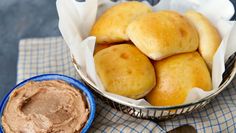 a basket filled with rolls next to a bowl of peanut butter and a spoon on a blue checkered cloth