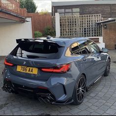 the rear end of a grey car parked in front of a brick driveway with two cars behind it