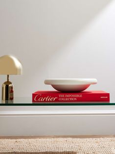 a glass table topped with two books and a bowl on top of each other next to a lamp