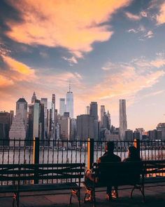 two people are sitting on a bench in front of the cityscape at sunset