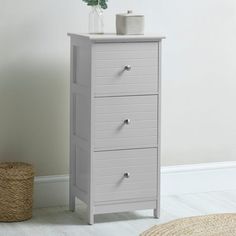 a white cabinet with three drawers next to a rug and vase on the floor in front of it