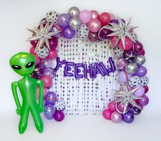 a green alien standing in front of a frame with balloons and star decorations on it