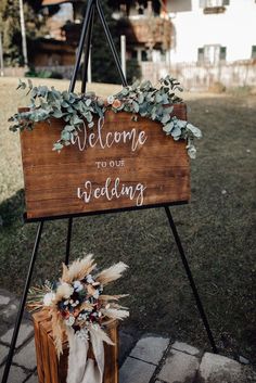 a welcome to our wedding sign with greenery and feathers on the bottom, sitting in front of a house