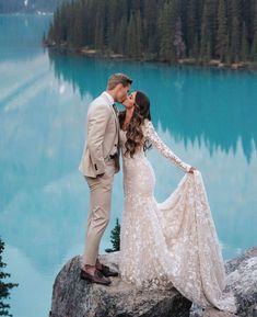 a bride and groom kissing on the edge of a cliff overlooking a lake with mountains in the background