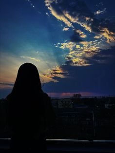 a woman standing in front of a window with the sun setting behind her and clouds