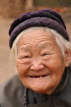 an old woman with white hair and a purple hat smiles at the camera while wearing a black jacket