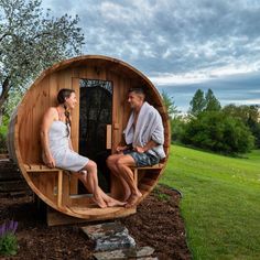 a man and woman sitting in a wooden barrel sauna with the door open, outside