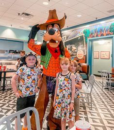 two children standing in front of goofy the dog mascot at an indoor restaurant with other kids