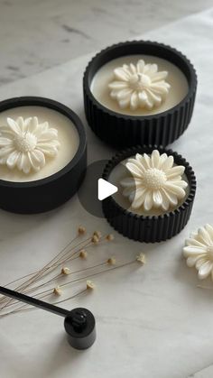 three black containers filled with white flowers on top of a marble counter next to a spoon