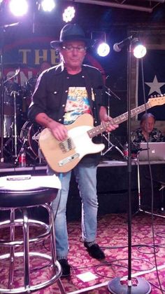 a man standing on top of a stage with a guitar in front of him and two microphones behind him