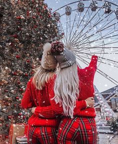 two women in matching red and black pajamas standing next to a christmas tree with a ferris wheel behind them