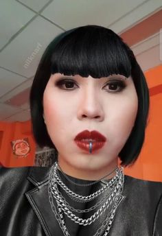 a woman with black hair and piercings on her nose wearing a leather jacket, silver chain necklace and red lipstick