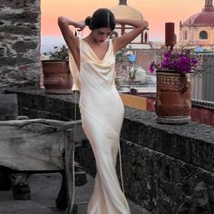 a woman in a long white dress is standing on a ledge with her hands behind her head