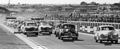 vintage cars driving on a race track with spectators in the background