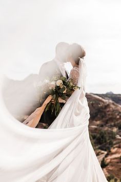 a bride and groom kissing on top of a mountain with their veil blowing in the wind