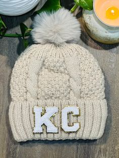 a white knit hat with the letter k c on it and a candle next to it