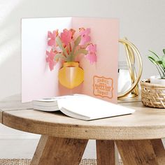 a card with pink flowers in a yellow vase on a table next to a potted plant