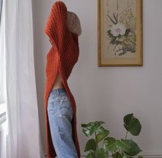 a woman standing in front of a window with her back turned to the camera, wearing an orange sweater and jeans