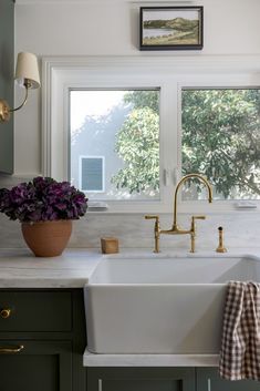 a white sink sitting under a window next to a potted purple flower on top of a counter