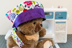 a brown teddy bear wearing a purple hat sitting next to a sewing machine on a table