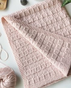 a pink knitted blanket next to some yarn and knitting needles on a white surface