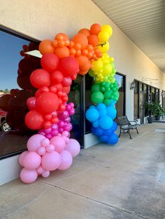 a rainbow colored balloon arch on the side of a building with a bench in front of it