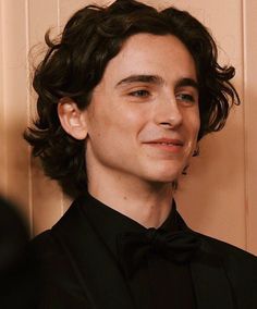 a young man in a black shirt and bow tie looking at the camera while standing next to a wall