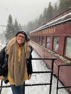 a woman standing in front of a red train car on snow covered ground with trees behind her