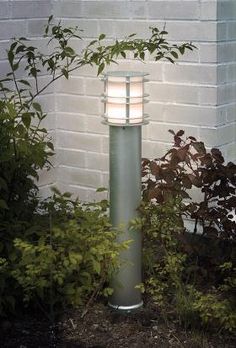 a lamp that is sitting in the grass near some bushes and trees, next to a brick wall