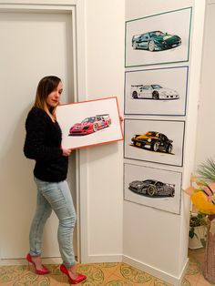 a woman standing next to a wall holding up a framed poster with cars on it