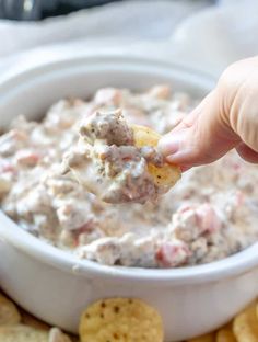 a hand holding a cracker over a bowl of dip with chips on the side